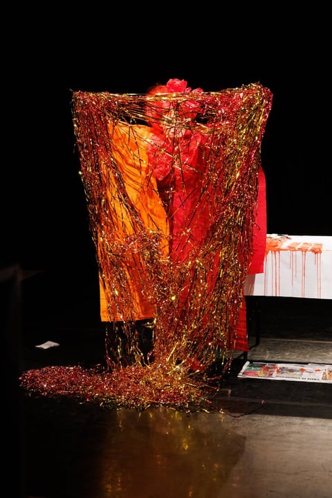 A chair draped in cascading red-and-gold tinsel over orange fabric, pooling across the dark stage floor like a glittering waterfall.