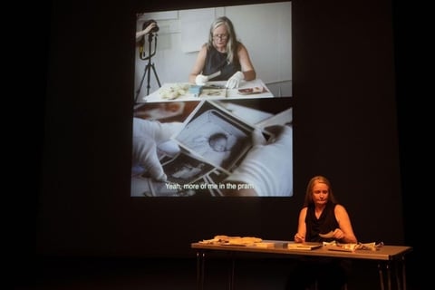 On a darkened stage, Sue MacLaine sits at a small table handling papers, while a large projection behind her shows white-gloved hands examining old photographs, including a framed baby portrait captioned 'Yeah, more of me in the pram'.