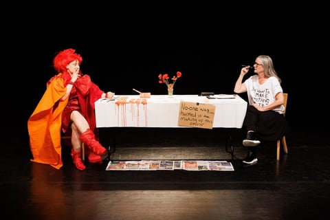 Sue MacLaine and Hannah Ringham face each other across a white-clothed table set with orange flowers and trailing red paint; a cardboard sign reads 'No-one was harmed in the making of this show'.