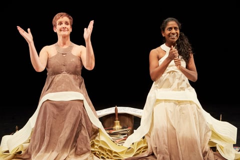 Sue MacLaine and Nadia Nadarajah kneel in their spreading paper gowns, smiling as they gesture — Sue with palms raised, Nadia with hands held together.