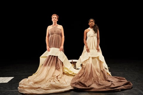 Sue MacLaine and Nadia Nadarajah stand facing forward in full-length dip-dyed gowns whose long paper trains spread across the stage, each with a printed text panel hanging at the front.