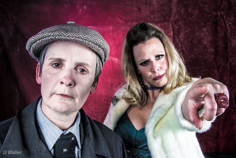 Against a dark-red velvet backdrop, Sue MacLaine as Sid Lester wears a tweed flat cap, suit and tie, while Valerie, in a white fur stole and teal dress, points forward wearing a large red ring.