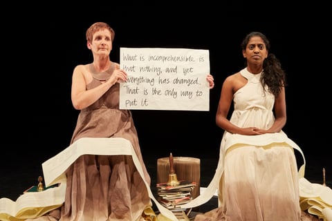 Two performers from Can I Start Again Please seated amid cascading book pages on a dark stage. Sue MacLaine, in a brown dress, holds a handwritten placard reading 'What is incomprehensible is that nothing, and yet everything has changed. That is the only way to put it', beside Nadia Nadarajah in a cream dress.