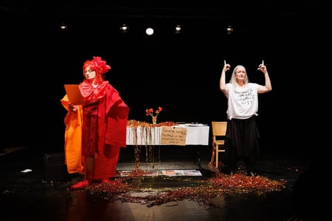 On a black stage scattered with red tinsel, Hannah Ringham stands in her red costume holding a board while Sue MacLaine, in her slogan t-shirt, raises both arms with index fingers pointing upward.
